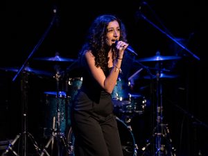 Veronica Mansour, microphone in hand, sings on stage. A drum set is behind her.